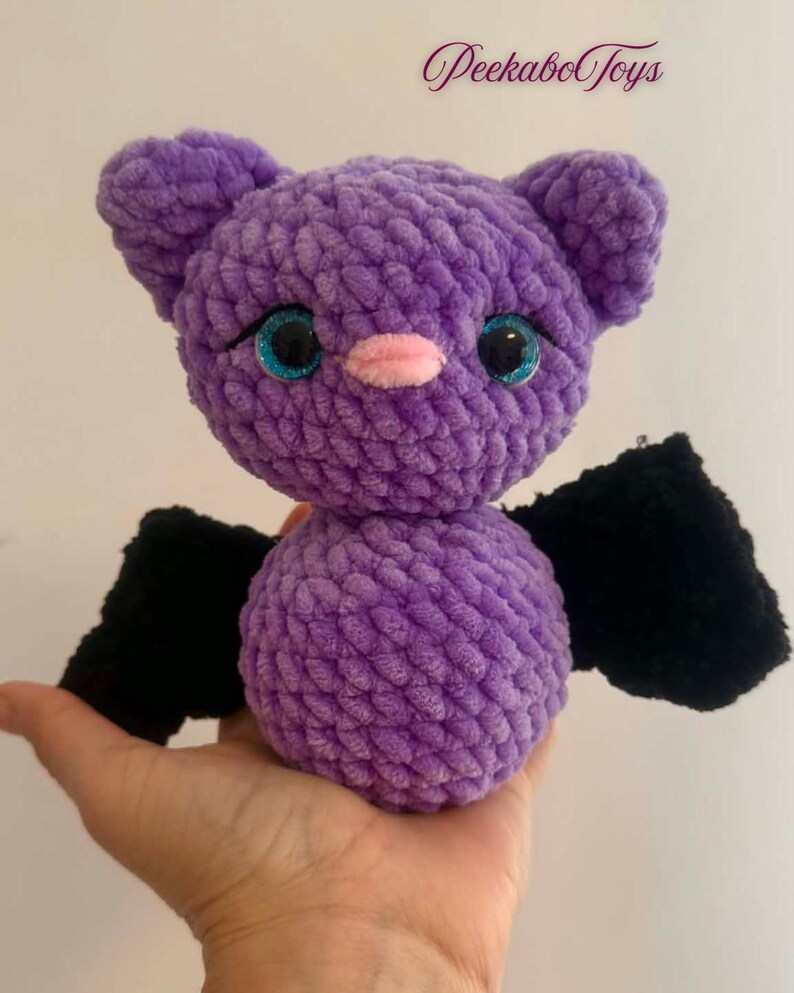 May include: A handmade purple crochet bat plush toy with black wings and sparkly blue eyes. The bat has a pink nose and small ears. The text "PeekaboToys" is in the upper right corner.