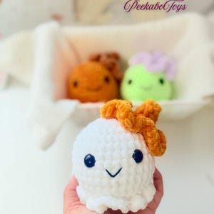 May include: A white crocheted plush toy with a smiling face and orange bow. Two other plush toys are in the background, one orange and one green with a purple top. The toys are in a white basket. The text "PeekaboToys" is in the upper right corner.