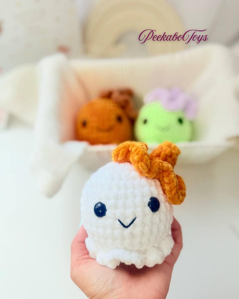 May include: A white crocheted plush toy with a smiling face and orange bow. Two other plush toys are in the background, one orange and one green with a purple top. The toys are in a white basket. The text "PeekaboToys" is in the upper right corner.