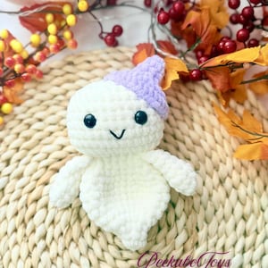 May include: A handmade white crochet ghost toy with a lavender hat, black eyes, and a smiling mouth. The ghost has small arms and a rounded body. The text "Peekaboo Toys" is visible in the bottom right corner. The toy is placed on a woven mat with fall foliage.