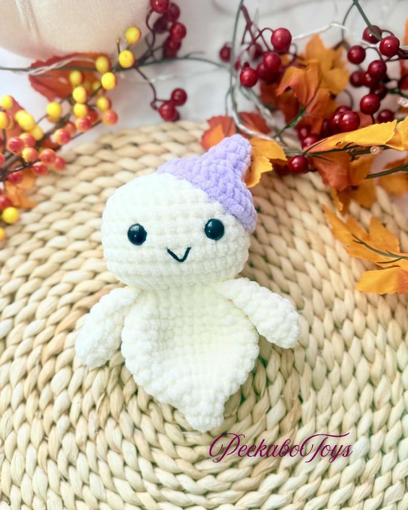 May include: A handmade white crochet ghost toy with a lavender hat, black eyes, and a smiling mouth. The ghost has small arms and a rounded body. The toy is sitting on a woven mat with fall foliage in the background. The text "Peekaboo Toys" is visible.