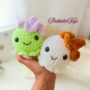 May include: Two crocheted plush toys. One is light green with a lavender flower-like top, the other is white with an orange top. Both have black eyes and smiling faces. The text "Peekabo Toys" is in the background.