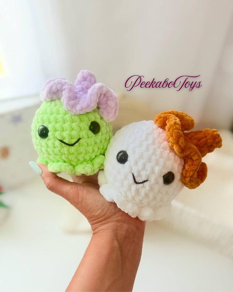 May include: Two crocheted plush toys. One is light green with a lavender flower-like top, the other is white with an orange top. Both have black eyes and smiling faces. The text "Peekabo Toys" is in the background.