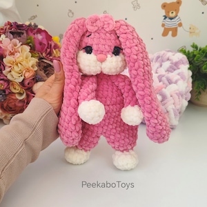 May include: A plush pink bunny toy with long floppy ears, white accents, and a sweet expression. The bunny is wearing a pink jumpsuit and has white paws. The text "PeekaboToys" is visible on the surface.