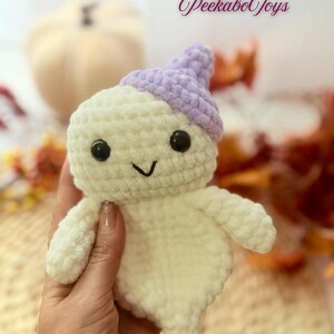May include: A handmade crochet ghost toy in off-white with a lavender hat. The ghost has black safety eyes and a stitched smile. The text "Peekabo Toys" is in the upper right corner. The toy is held in a hand.