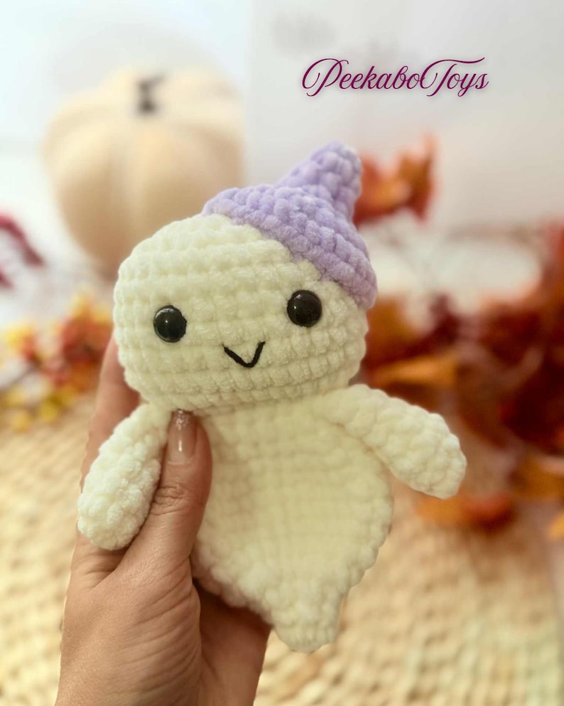 May include: A handmade crochet ghost toy in off-white with a lavender hat. The ghost has black safety eyes and a stitched smile. The text "Peekabo Toys" is in the upper right corner. The toy is held in a hand.