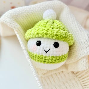 May include: A small crocheted snowman toy wearing a green hat with a white pom-pom. The snowman has a white body with black eyes and a small pink nose.