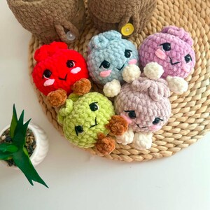 Puede incluir: Una colecci&oacute;n de seis peque&ntilde;as figuras de peluche tejidas a ganchillo, redondas, con rasgos faciales bordados, incluyendo ojos negros y mejillas rosadas. Las figuras son rojas, azul claro, moradas, verde claro y lavanda, junto a dos recipientes marrones.