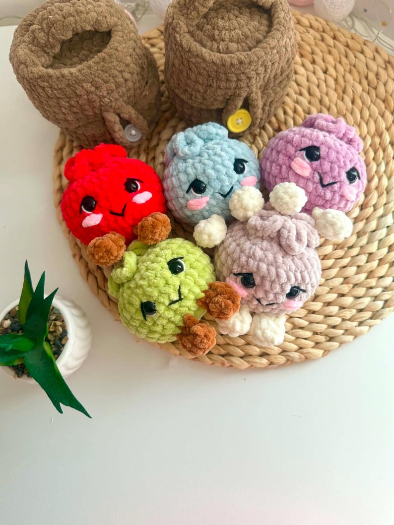Puede incluir: Una colecci&oacute;n de seis peque&ntilde;as figuras de peluche tejidas a ganchillo, redondas, con rasgos faciales bordados, incluyendo ojos negros y mejillas rosadas. Las figuras son rojas, azul claro, moradas, verde claro y lavanda, junto a dos recipientes marrones.