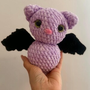 May include: A handmade lavender crochet bat plush toy with black wings, large googly eyes, and a pink nose. The toy is held in a hand and features the text "PeekaboToys" in the upper right corner.