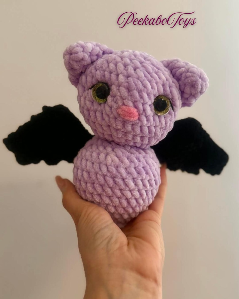 May include: A handmade lavender crochet bat plush toy with black wings, large googly eyes, and a pink nose. The toy is held in a hand and features the text "PeekaboToys" in the upper right corner.