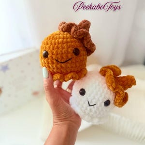 May include: Two handmade crochet plush toys. One is orange, the other is white. Both have brown accents and smiling faces with black eyes. The toys are held in a hand. The text "PeekaboToys" is in the background.