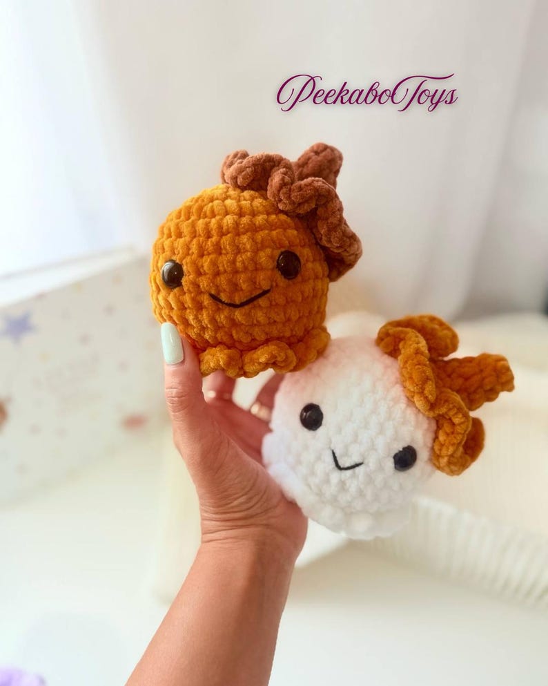 May include: Two handmade crochet plush toys. One is orange, the other is white. Both have brown accents and smiling faces with black eyes. The toys are held in a hand. The text "PeekaboToys" is in the background.