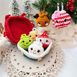 May include: A collection of crocheted plush ornaments featuring round, smiling faces with pink cheeks. The set includes a green, tan, red-and-white striped, and white ornament, all nestled in a dark green and white crocheted basket.