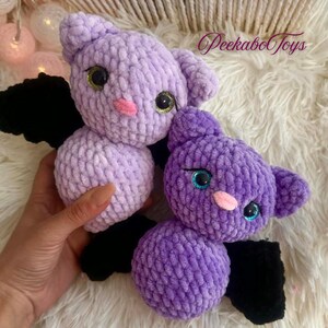 May include: Two handmade crochet bats in shades of purple. The bats have black wings, pink noses, and large eyes with gold and blue accents. The text "Peekabo Toys" is visible in the upper right corner.
