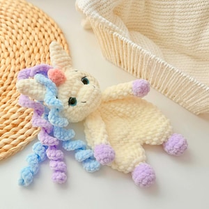 May include: A handmade crochet unicorn plush toy in cream, with blue and purple mane, and lavender hooves. The unicorn has blue eyes and a pink horn. The plush is lying on a white surface, with a woven mat and a white basket in the background.