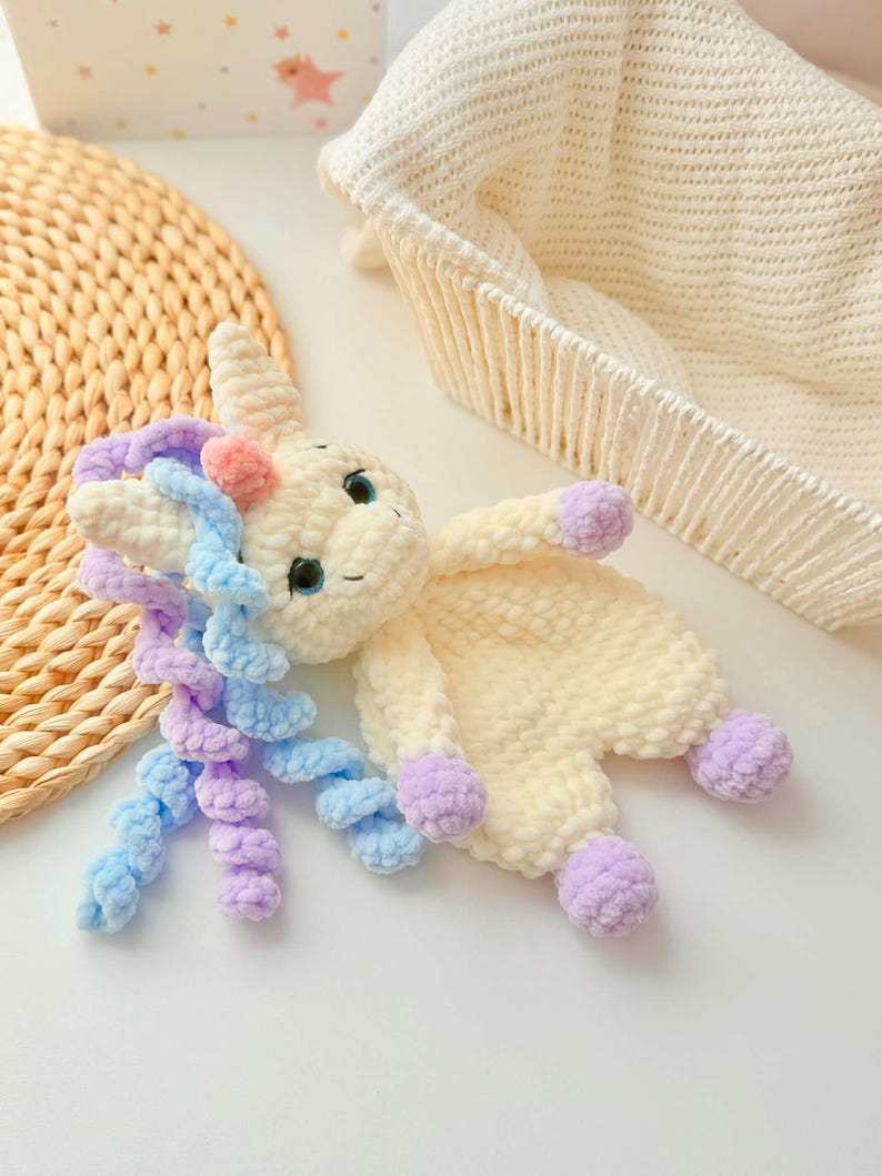 May include: A handmade crochet unicorn plush toy in cream, with blue and purple mane, and lavender hooves. The unicorn has blue eyes and a pink horn. The plush is lying on a white surface, with a woven mat and a white basket in the background.
