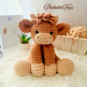 Può includere: Un peluche a forma di mucca fatto a mano all'uncinetto, nei toni del marrone e del beige. La mucca di peluche ha grandi orecchie, una testa soffice e un viso sorridente. Il testo "Peekabo Toys" è nell'angolo in alto a destra. Il giocattolo è su una superficie bianca e soffice.