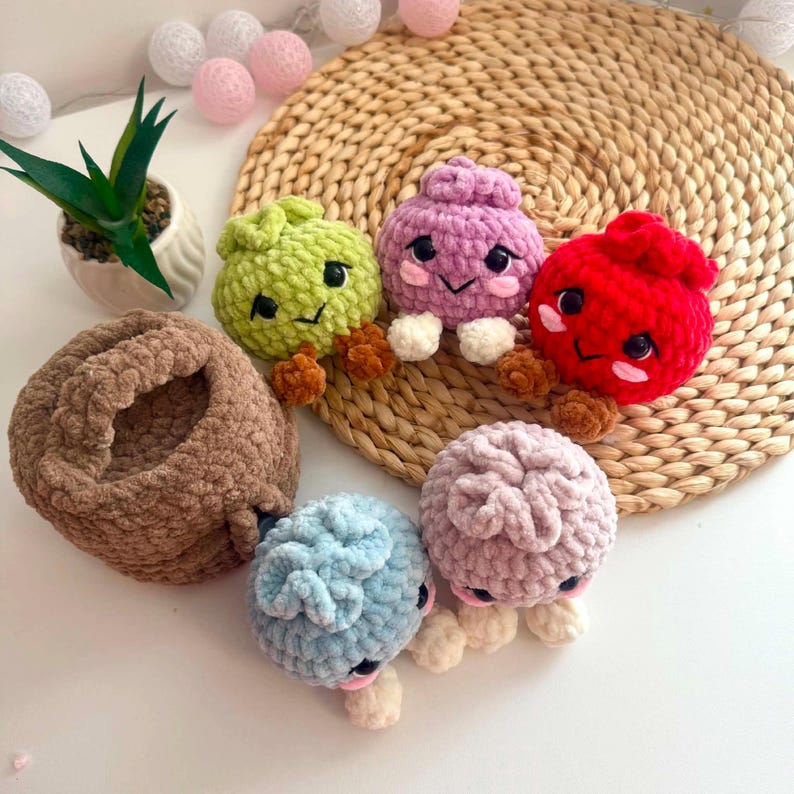 Puede incluir: Un conjunto de cinco peque&ntilde;as criaturas redondas de ganchillo afelpado en verde lima, lavanda, rojo brillante, azul claro y rosa empolvado, cada una con ojos de seguridad negros, sonrisas bordadas y peque&ntilde;os pies marrones o crema.