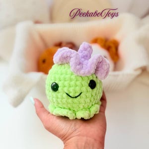 May include: A handmade green crochet toy with a smiling face and a lavender flower-like detail on top. The toy is held in a hand, with the text "PeekaboToys" visible in the background. The toy is a cute, whimsical design.