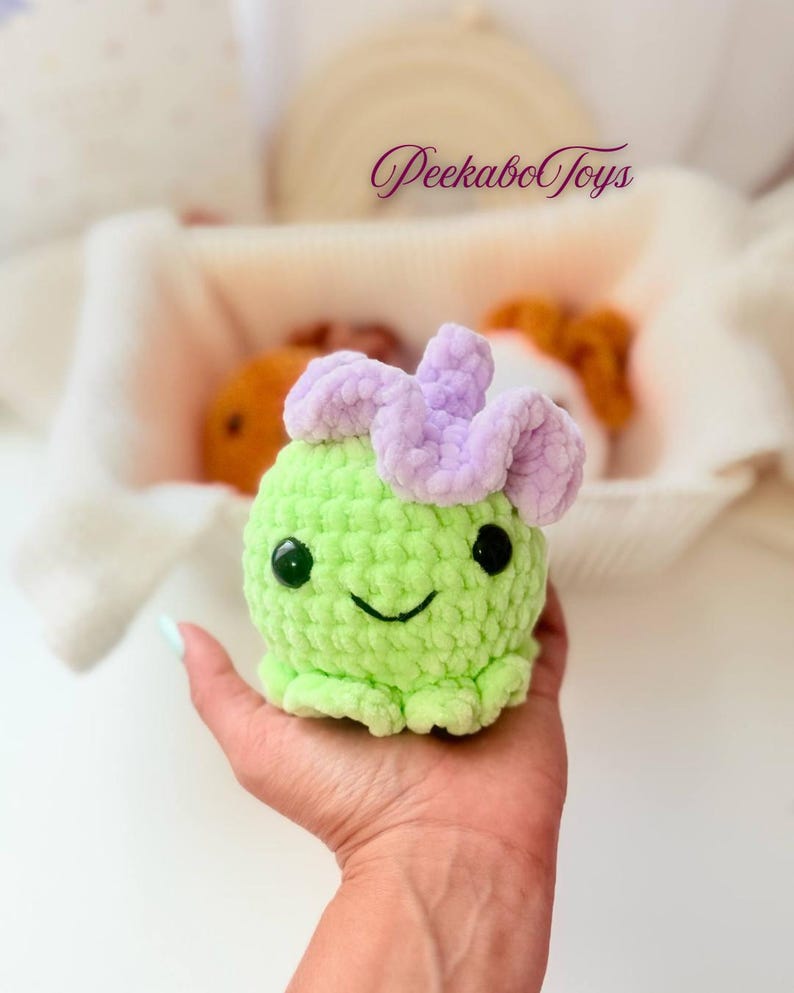 May include: A handmade green crochet toy with a smiling face and a lavender flower-like detail on top. The toy is held in a hand, with the text "PeekaboToys" visible in the background. The toy is a cute, whimsical design.