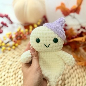 May include: A handmade, plush ghost toy with a light purple hat and black button eyes. The ghost is off-white and has a smiling face. The text "Peekabo Toys" is in the upper right corner. The toy is held in a hand.