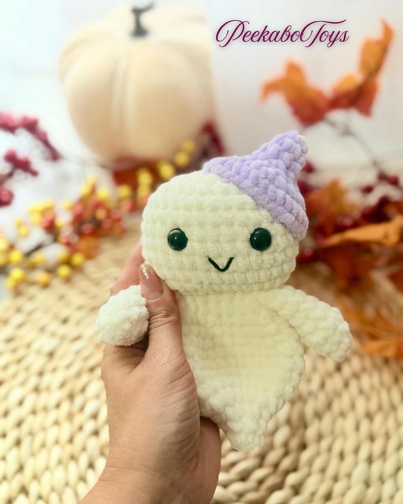 May include: A handmade, plush ghost toy with a light purple hat and black button eyes. The ghost is off-white and has a smiling face. The text "Peekabo Toys" is in the upper right corner. The toy is held in a hand.