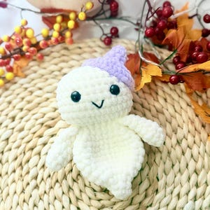 May include: A handmade, white crochet ghost with a lavender hat, black eyes, and a stitched smile. The ghost is a plush toy, perfect for Halloween or as a decorative item. The background includes fall foliage and the text "Peekabo Toys".