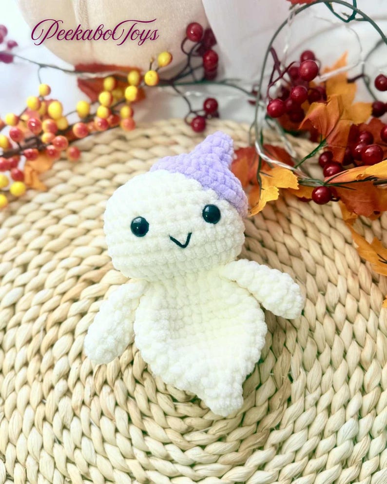 May include: A handmade, white crochet ghost with a lavender hat, black eyes, and a stitched smile. The ghost is a plush toy, perfect for Halloween or as a decorative item. The background includes fall foliage and the text "Peekabo Toys".