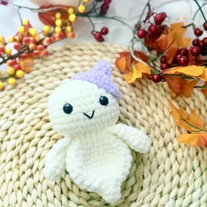 May include: A handmade white crochet ghost toy with a lavender hat and black eyes. The ghost has a smiling face and small arms. The text "PeekaboToys" is in the upper left corner. The ghost is on a woven mat with fall foliage.
