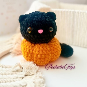 May include: A crocheted cat toy with a black head and tail, and an orange body. The cat has large, amber-coloured eyes and a small pink nose. The toy is made of soft, textured yarn, and is likely a children's toy.