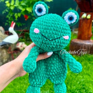 May include: A handmade green crochet frog plush toy with large blue and white eyes, pink cheeks, and a small pink tongue. The plush is held in front of a green grassy background. The text "PeekaboToys" is visible.