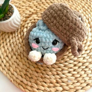 Puede incluir: Un peluche de ganchillo azul claro con cara sonriente, ojos negros y mejillas rosadas, metido dentro de una c&aacute;scara de ganchillo marr&oacute;n con forma de bellota. El juguete tiene dos peque&ntilde;as manos blancas de ganchillo y una tapa marr&oacute;n.