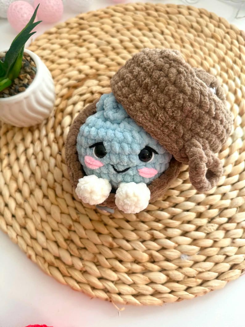 Puede incluir: Un peluche de ganchillo azul claro con cara sonriente, ojos negros y mejillas rosadas, metido dentro de una c&aacute;scara de ganchillo marr&oacute;n con forma de bellota. El juguete tiene dos peque&ntilde;as manos blancas de ganchillo y una tapa marr&oacute;n.