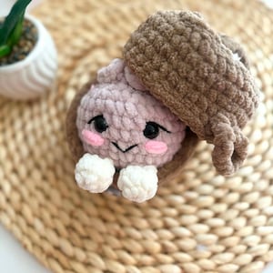 Puede incluir: Un juguete de ganchillo de felpa con forma de criatura rosa claro metida dentro de una c&aacute;scara de bellota marr&oacute;n. La criatura tiene ojos de seguridad negros, mejillas bordadas en rosa, una peque&ntilde;a sonrisa negra y dos manos blancas.