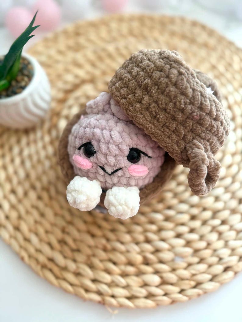 Puede incluir: Un juguete de ganchillo de felpa con forma de criatura rosa claro metida dentro de una c&aacute;scara de bellota marr&oacute;n. La criatura tiene ojos de seguridad negros, mejillas bordadas en rosa, una peque&ntilde;a sonrisa negra y dos manos blancas.