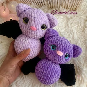 May include: Two handmade crochet bats in shades of purple. The bats have black wings, pink noses, and large eyes with gold and blue accents. The text "PeekaboToys" is visible in the upper right corner.