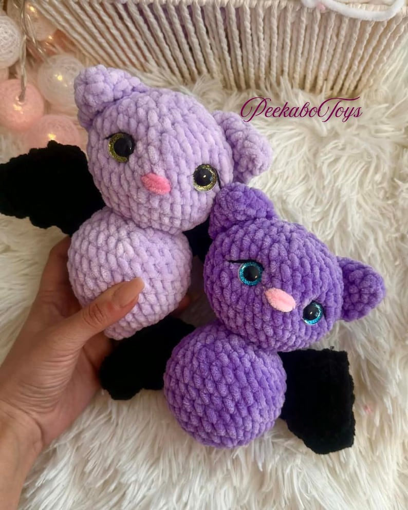 May include: Two handmade crochet bats in shades of purple. The bats have black wings, pink noses, and large eyes with gold and blue accents. The text "PeekaboToys" is visible in the upper right corner.
