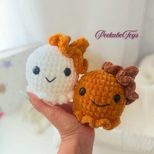 May include: Two crocheted plush toys, one white and one orange, each with a smiling face and a decorative top. The white toy has an orange top, and the orange toy has a brown top. The toys are held in a hand.