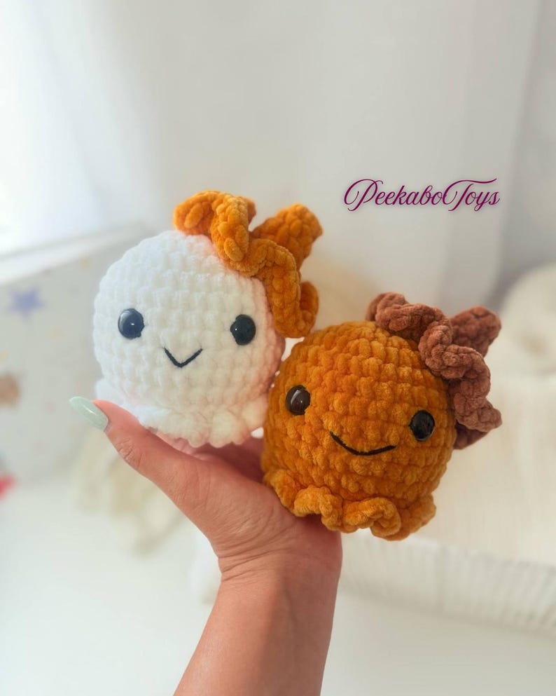 May include: Two crocheted plush toys, one white and one orange, each with a smiling face and a decorative top. The white toy has an orange top, and the orange toy has a brown top. The toys are held in a hand.