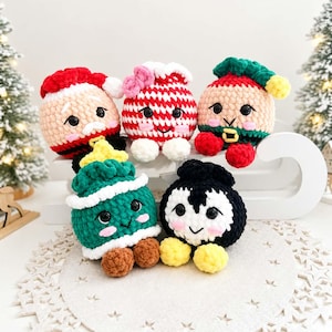 May include: Five round, plush, crocheted ornaments featuring character designs: a Santa Claus, a red and white striped peppermint, an elf, a green Christmas tree with a yellow star, and a black and white penguin, all with black eyes and pink cheeks.