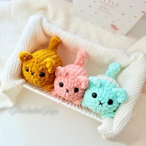 May include: Three crocheted plush cat toys in a white blanket. The cats are yellow, pink, and light blue. The cats have black eyes and a stitched-on nose and mouth.