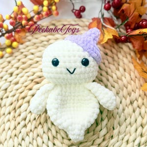 May include: A handmade white crochet ghost toy with a lavender hat. The ghost has black safety eyes and a stitched smile. The toy is on a woven mat with fall foliage in the background. The text "PeekaboToys" is visible.