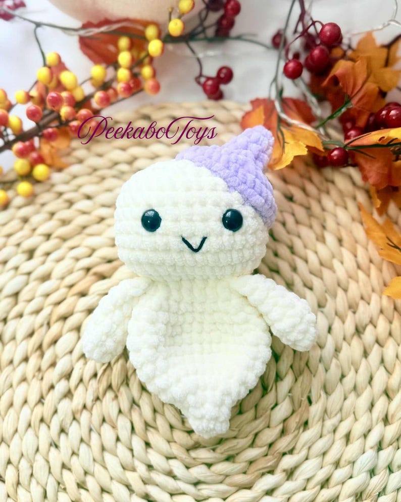 May include: A handmade white crochet ghost toy with a lavender hat. The ghost has black safety eyes and a stitched smile. The toy is on a woven mat with fall foliage in the background. The text "PeekaboToys" is visible.