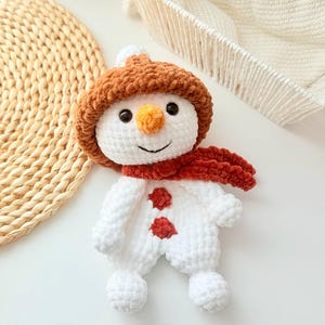 May include: A white crocheted snowman toy with a brown fuzzy hat and a red scarf. The snowman has two red buttons and a carrot nose.