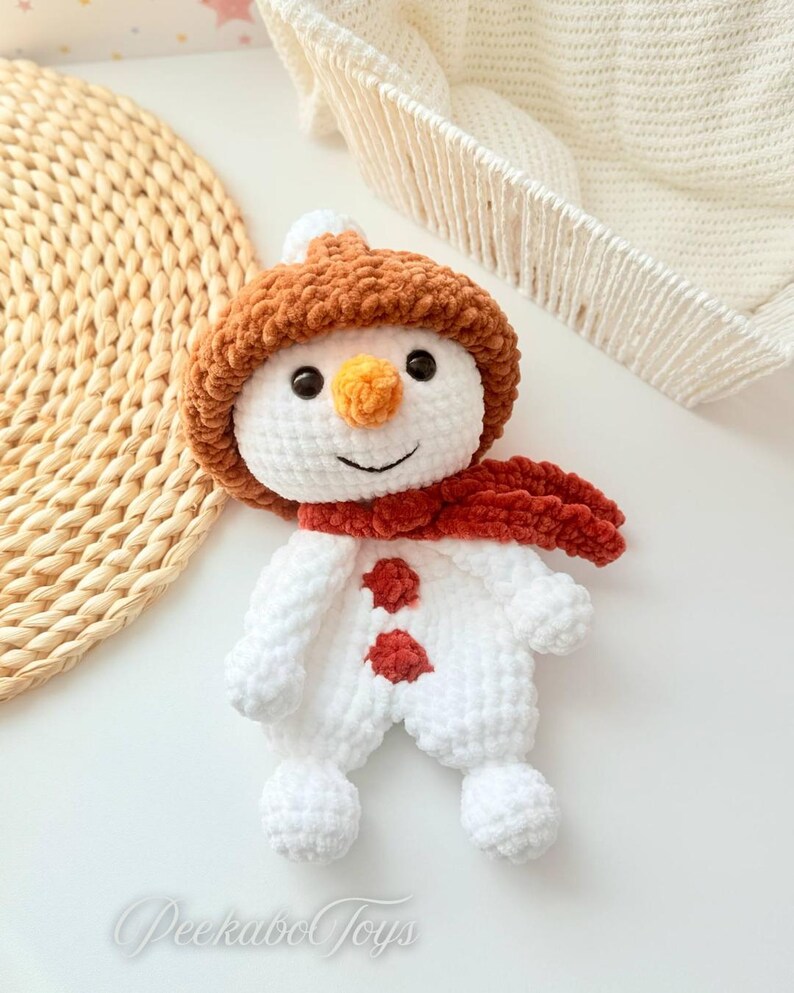 May include: A white crocheted snowman toy with a brown fuzzy hat and a red scarf. The snowman has two red buttons and a carrot nose.