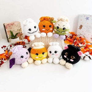 May include: A collection of seven small, round, crocheted plush figures with various facial expressions and colors, including white, orange, green, purple, yellow, and black. Each plush features small white feet and embroidered details.
