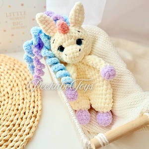 May include: A cream-colored crocheted unicorn toy with blue, purple, and pink accents. The unicorn has large, dark eyes and is lying in a white basket. The text "Peekaboo Toys" is visible on the toy.