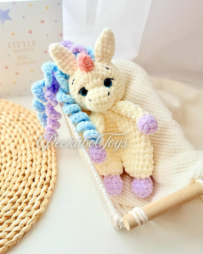 May include: A cream-colored crocheted unicorn toy with blue, purple, and pink accents. The unicorn has large, dark eyes and is lying in a white basket. The text "Peekaboo Toys" is visible on the toy.