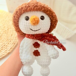 May include: A white crocheted snowman with a brown crocheted hat and a red scarf. The snowman has a carrot nose and black eyes. The snowman is holding a small brown crocheted heart.